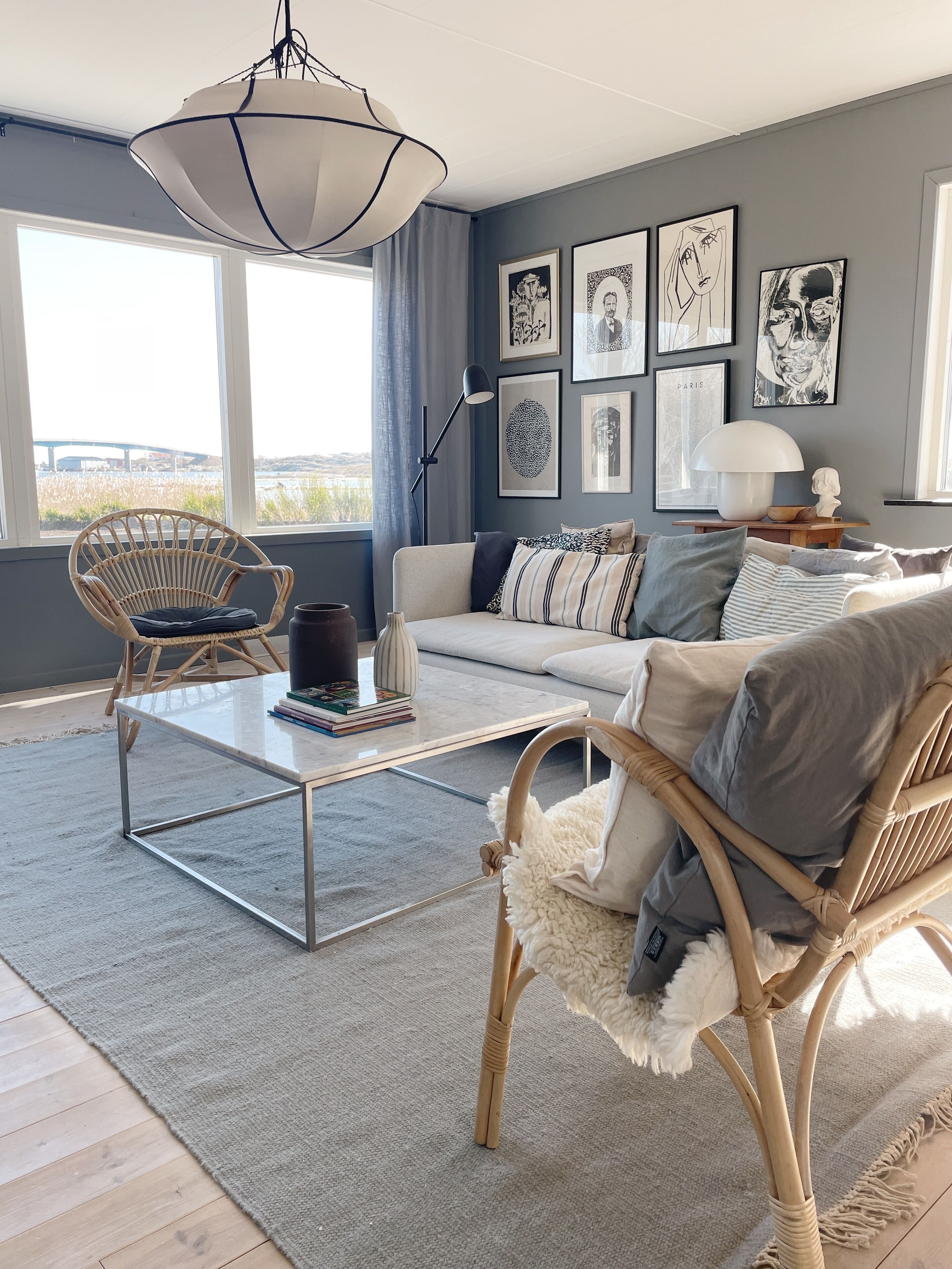 A modern living room with a sofa, two wicker chairs, a coffee table, and a large window offering a view of a bridge and fields.