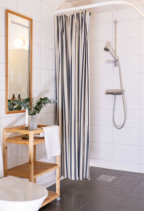 A bathroom with a wooden vanity, a mirror, a white sink, a shower with a striped curtain, and a potted plant. - Hönö Sjöbodar (Hono)