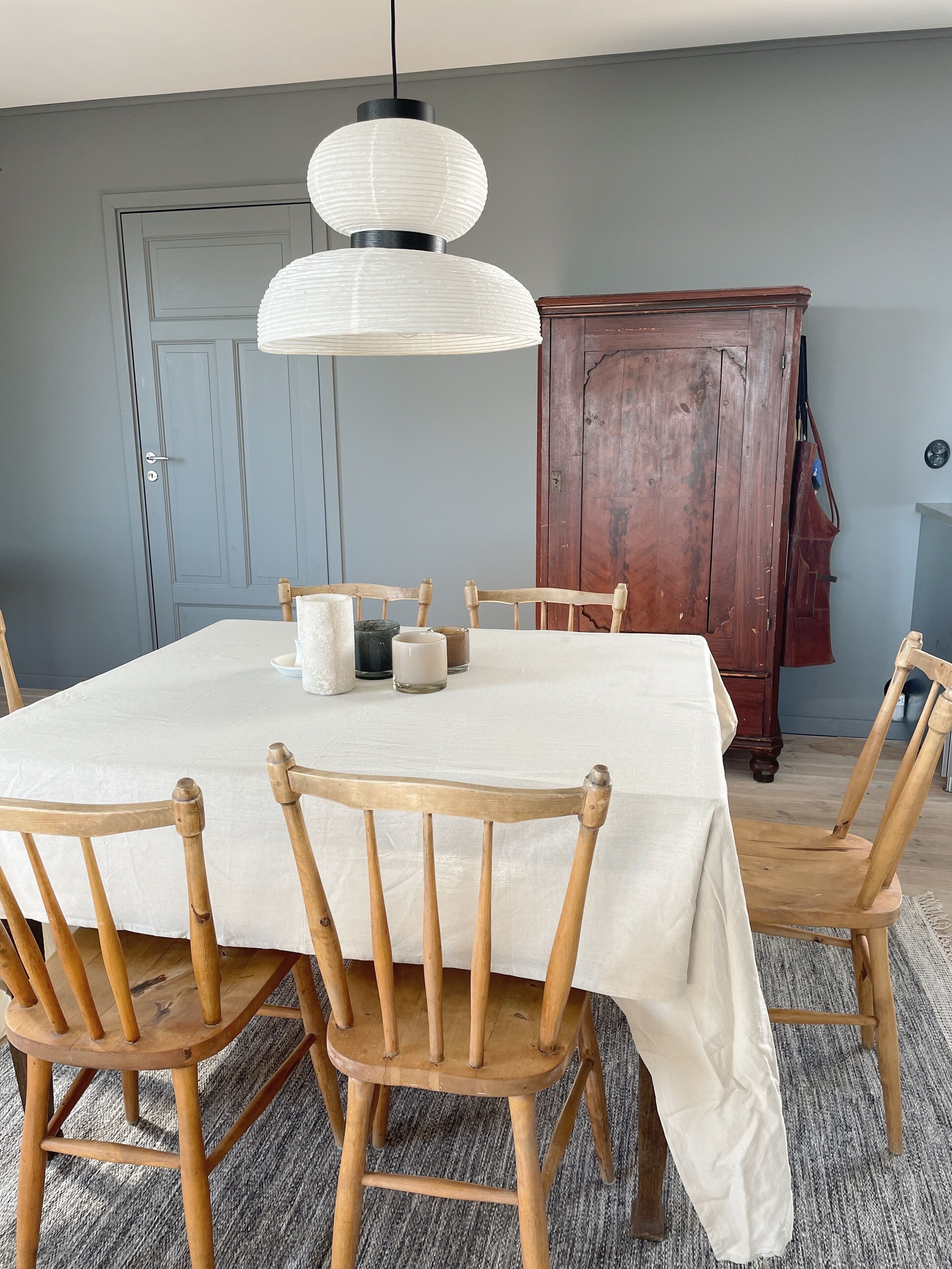 A dining area with a round table covered in a white tablecloth, surrounded by wooden chairs. A pendant light hangs above the table, and a wooden cabinet is in the background.