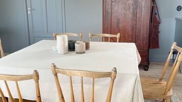 A dining area with a round table covered in a white tablecloth, surrounded by wooden chairs. A pendant light hangs above the table, and a wooden cabinet is in the background.