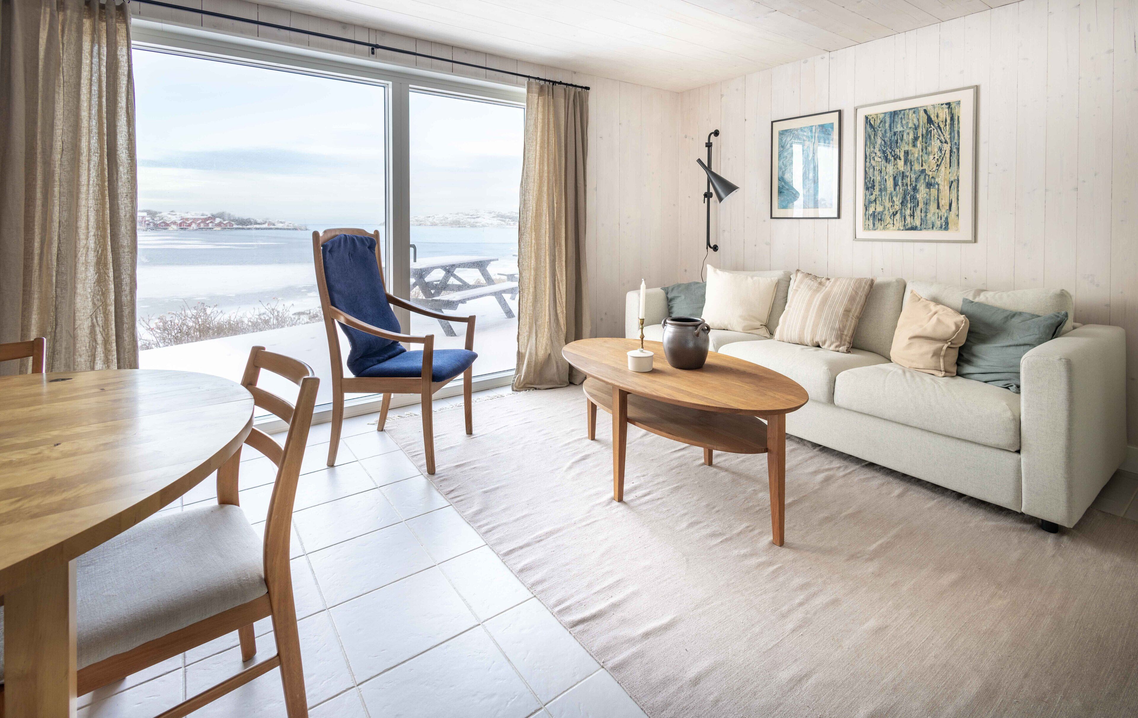 Uma sala de estar acolhedora com uma mesa de jantar de madeira, uma cadeira azul, um sofá branco com várias almofadas e vista para uma paisagem nevada através de grandes janelas.