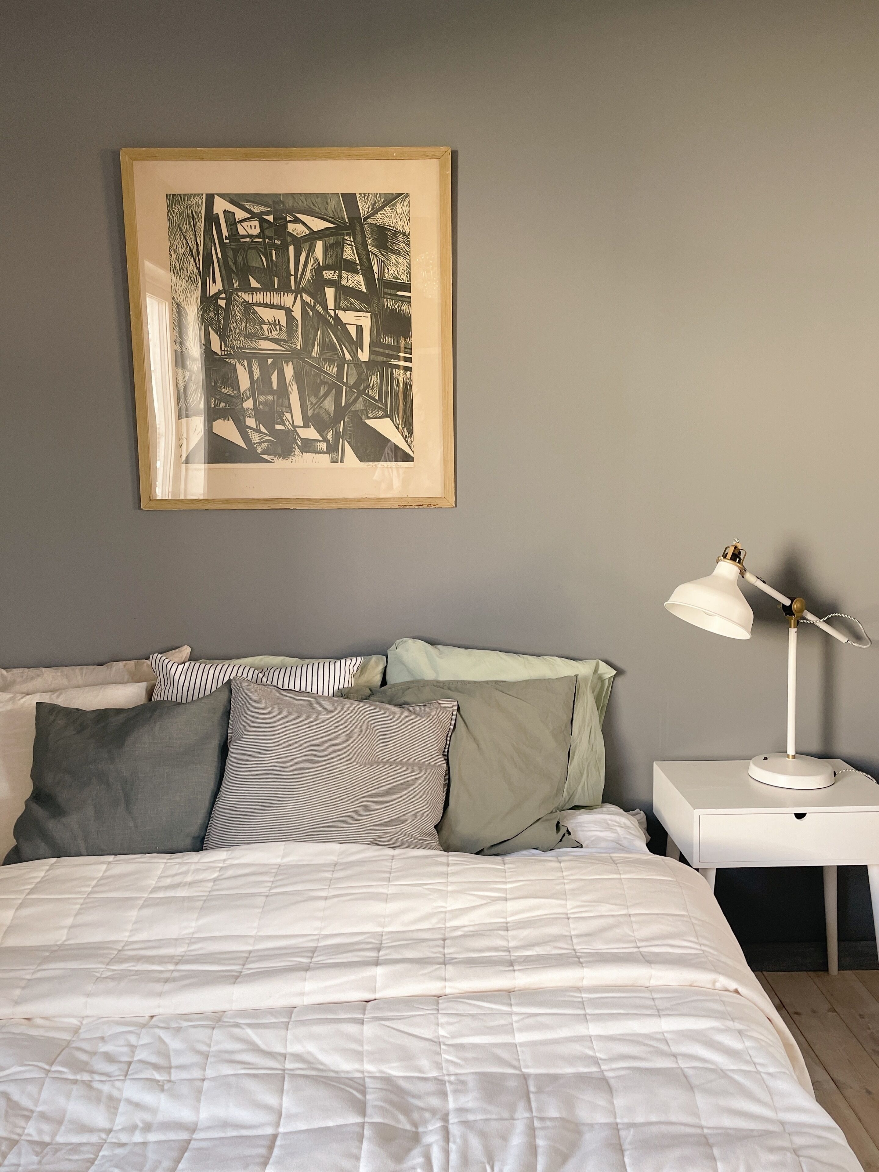 A neatly made bed with a white duvet, a bedside table with a lamp, and a framed abstract artwork on the wall.