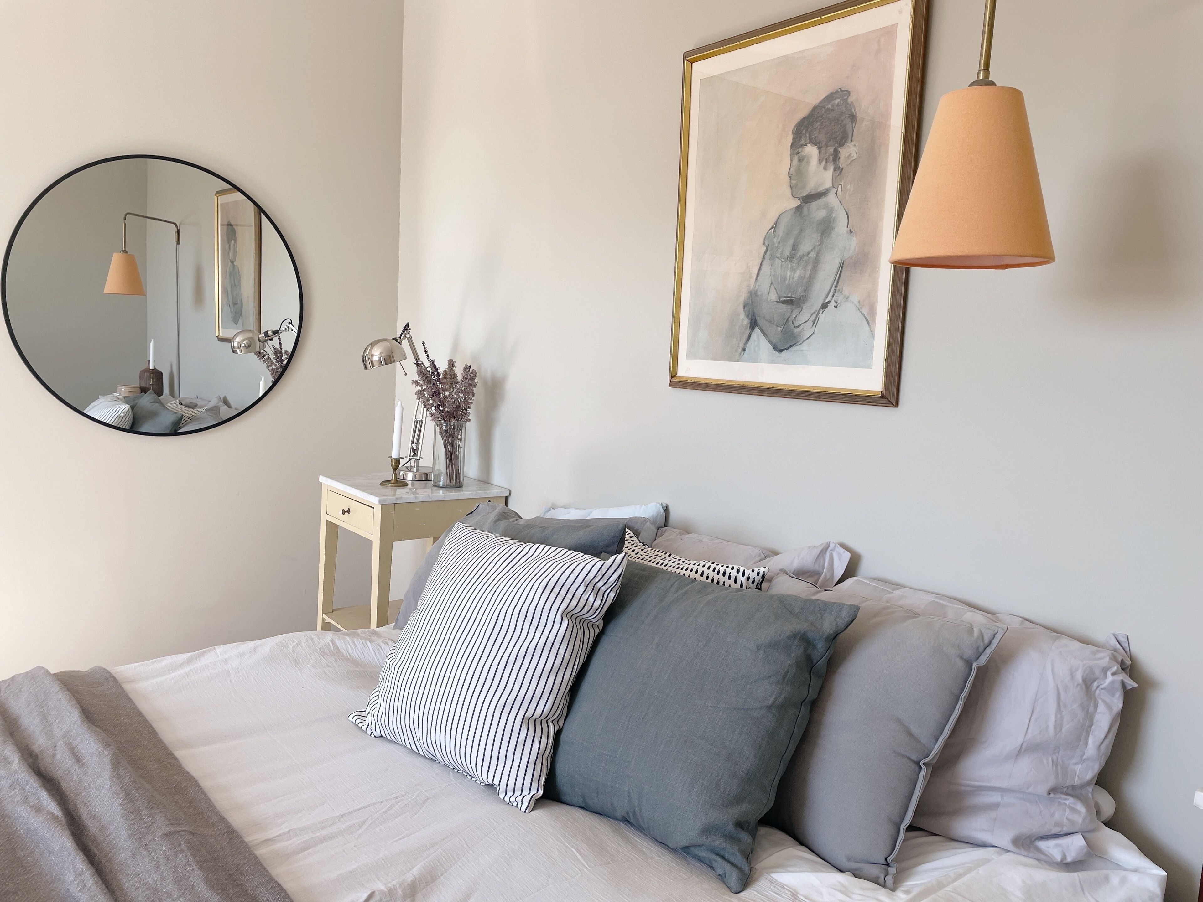 Un dormitorio ordenado con cama, almohadas, una mesita de noche, un espejo, una lámpara y una obra enmarcada en la pared.