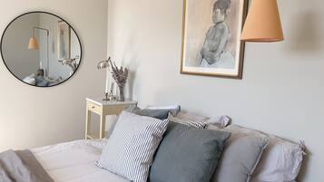 Un dormitorio ordenado con cama, almohadas, una mesita de noche, un espejo, una lámpara y una obra enmarcada en la pared.
