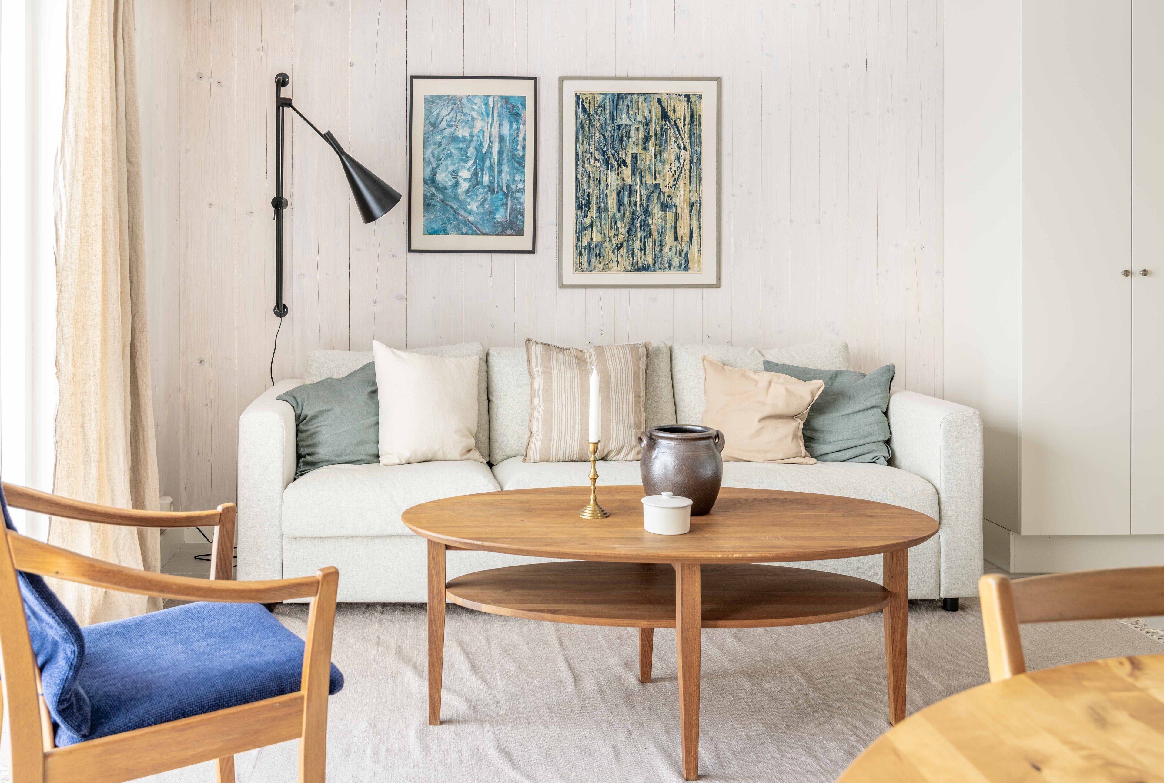 Una sala de estar con un sofá blanco, una mesa de centro redonda de madera y dos cuadros abstractos enmarcados en la pared.