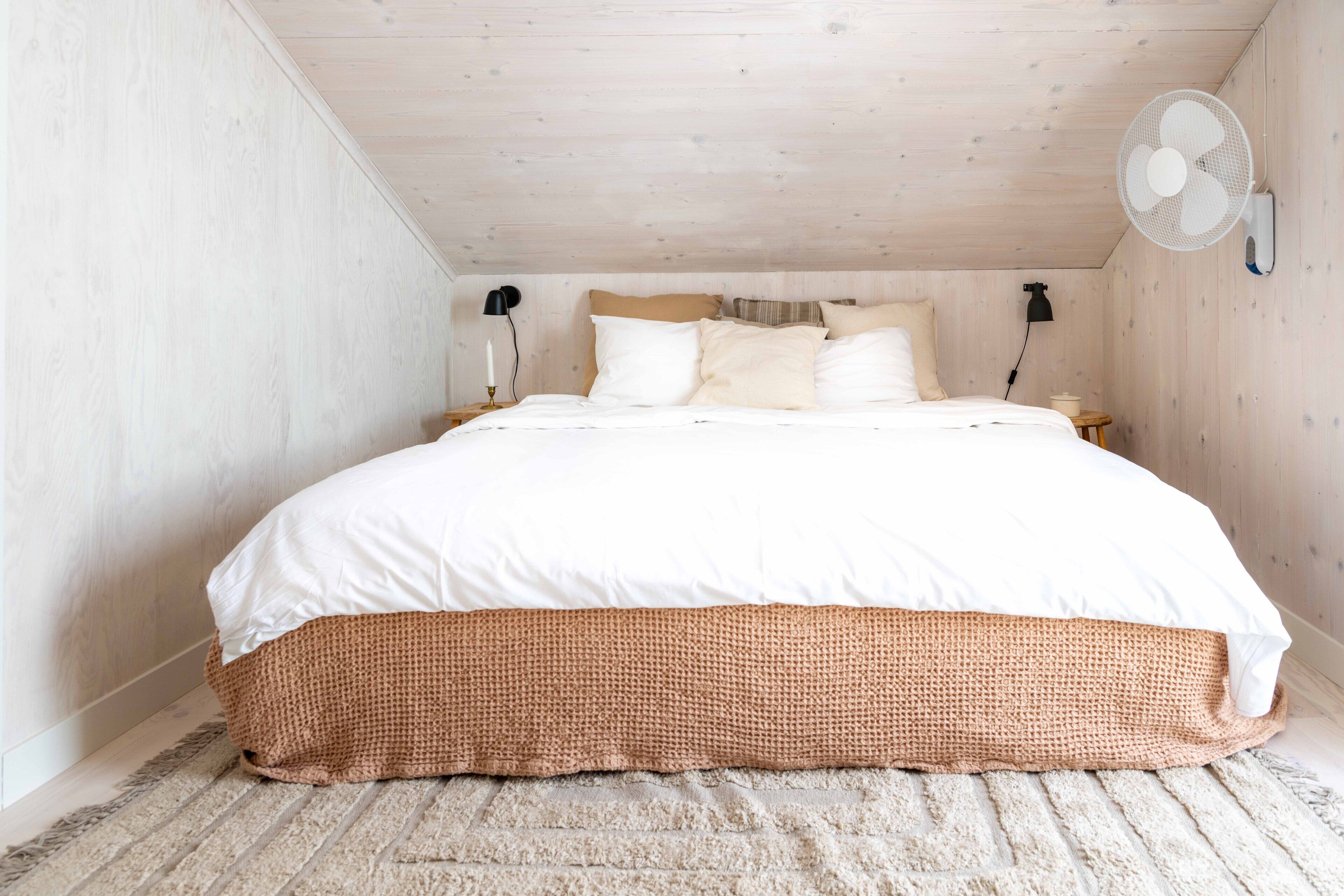 Een slaapkamer met een groot bed, een houten plafond, een ventilator aan de muur en een nachtkastje met een lamp.
