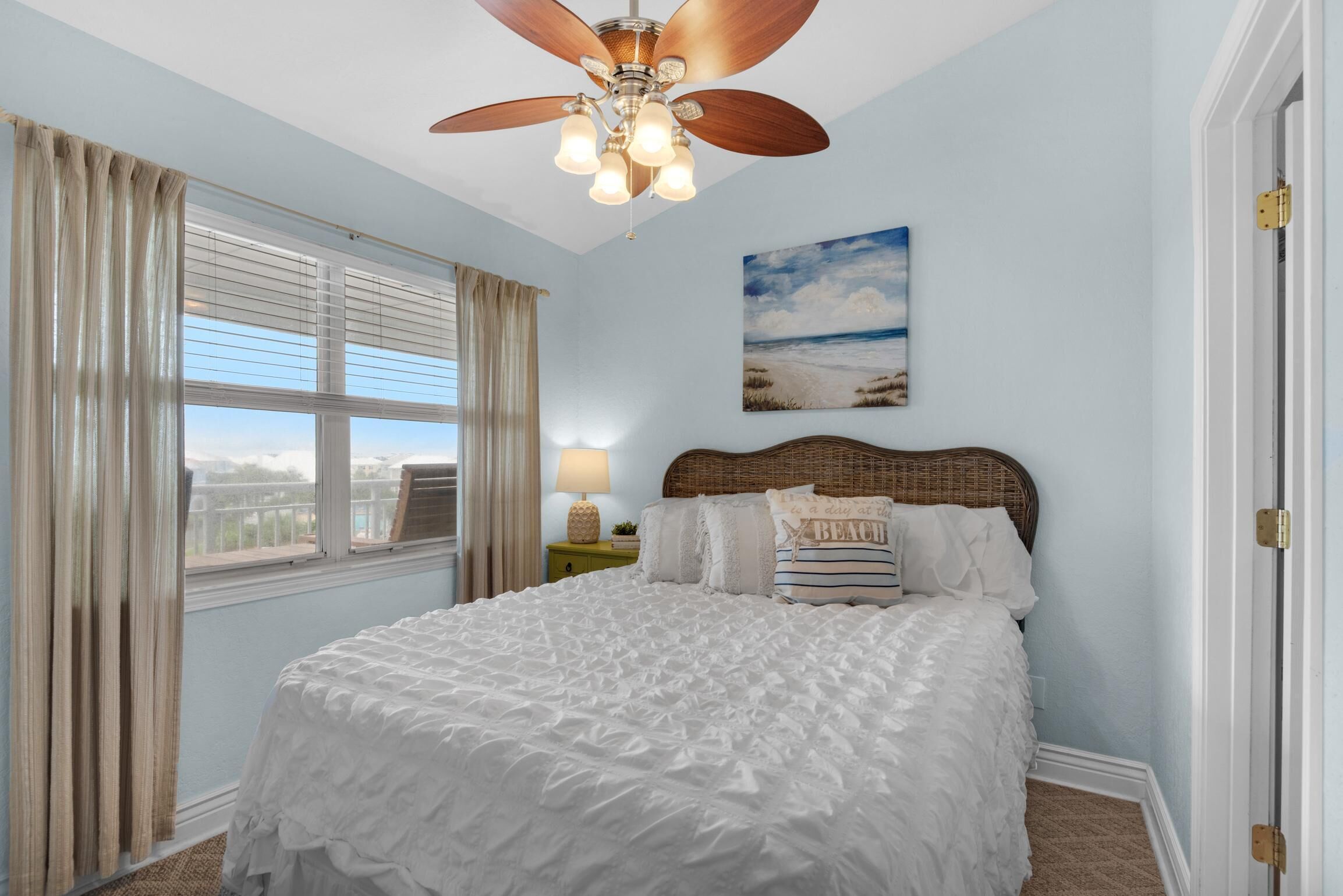 A bedroom with a large bed, a ceiling fan, a window with curtains, a nightstand with a lamp, and a beach-themed wall art.