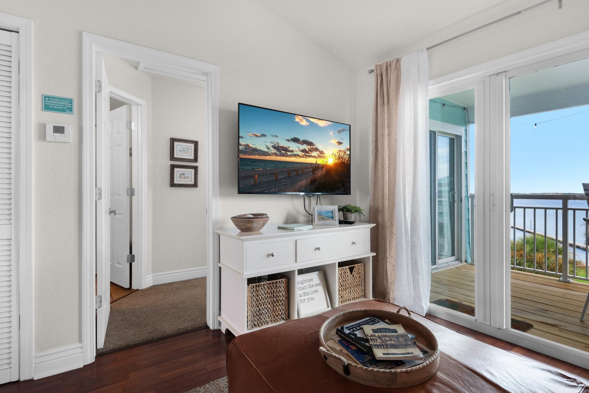 A room with a TV displaying a sunset over the ocean, a white console table with a bowl and a tray of books, and a large window with a view of a balcony.