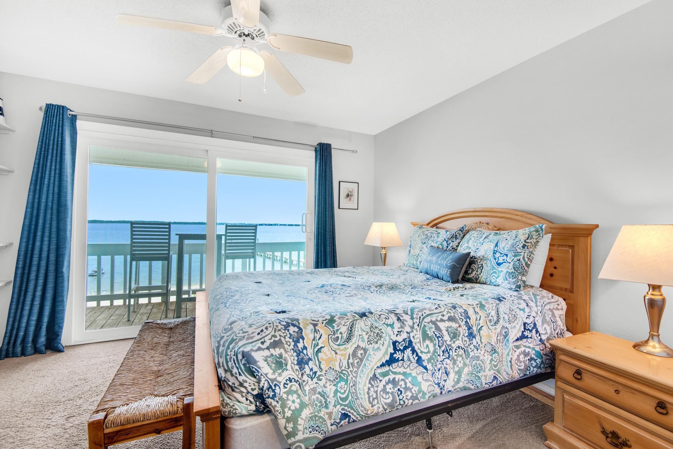 A bedroom with a large bed, a wooden nightstand, a ceiling fan, and a view of the ocean through a sliding glass door.