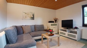 Ein Wohnzimmer mit einem grauen Ecksofa, einem Holztisch, einem Fernseher auf einem weißen Schrank und einem Gemälde an der Wand.