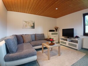 Ein Wohnzimmer mit einem grauen Ecksofa, einem Holztisch, einem Fernseher auf einem weißen Schrank und einem Gemälde an der Wand.