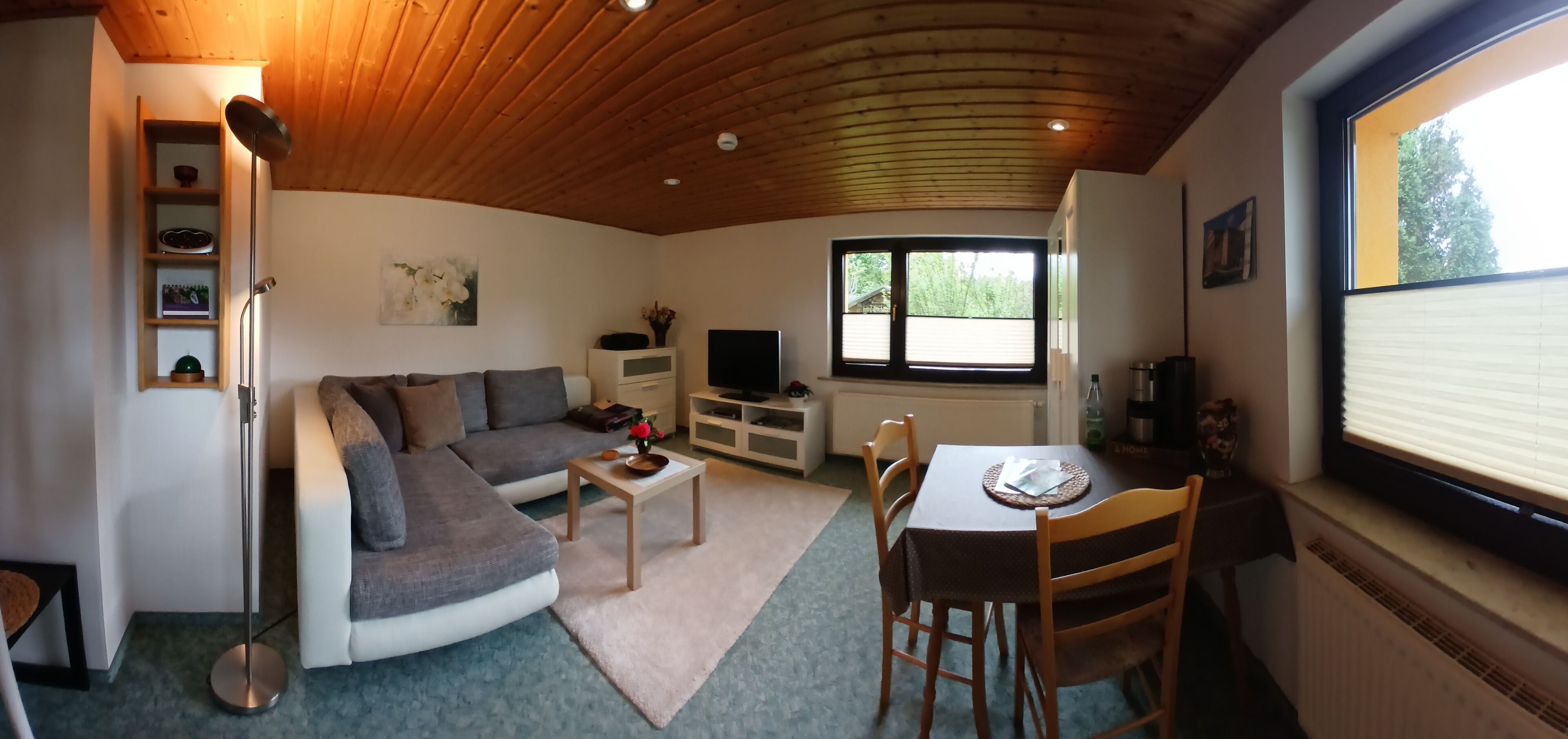 Ein gemütliches Wohnzimmer mit Sofa, Couchtisch und Esstisch. Ein Fernseher, ein Fenster mit Jalousien und eine Holzbalkendecke sind ebenfalls vorhanden.