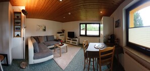 Ein gemütliches Wohnzimmer mit Sofa, Couchtisch und Esstisch. Ein Fernseher, ein Fenster mit Jalousien und eine Holzbalkendecke sind ebenfalls vorhanden.