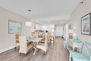 A modern kitchen with white cabinets, a dining area with wicker chairs, and a hallway with a lamp and picture frames. - Emerald Isle Unit 406 (Pensacola Beach)