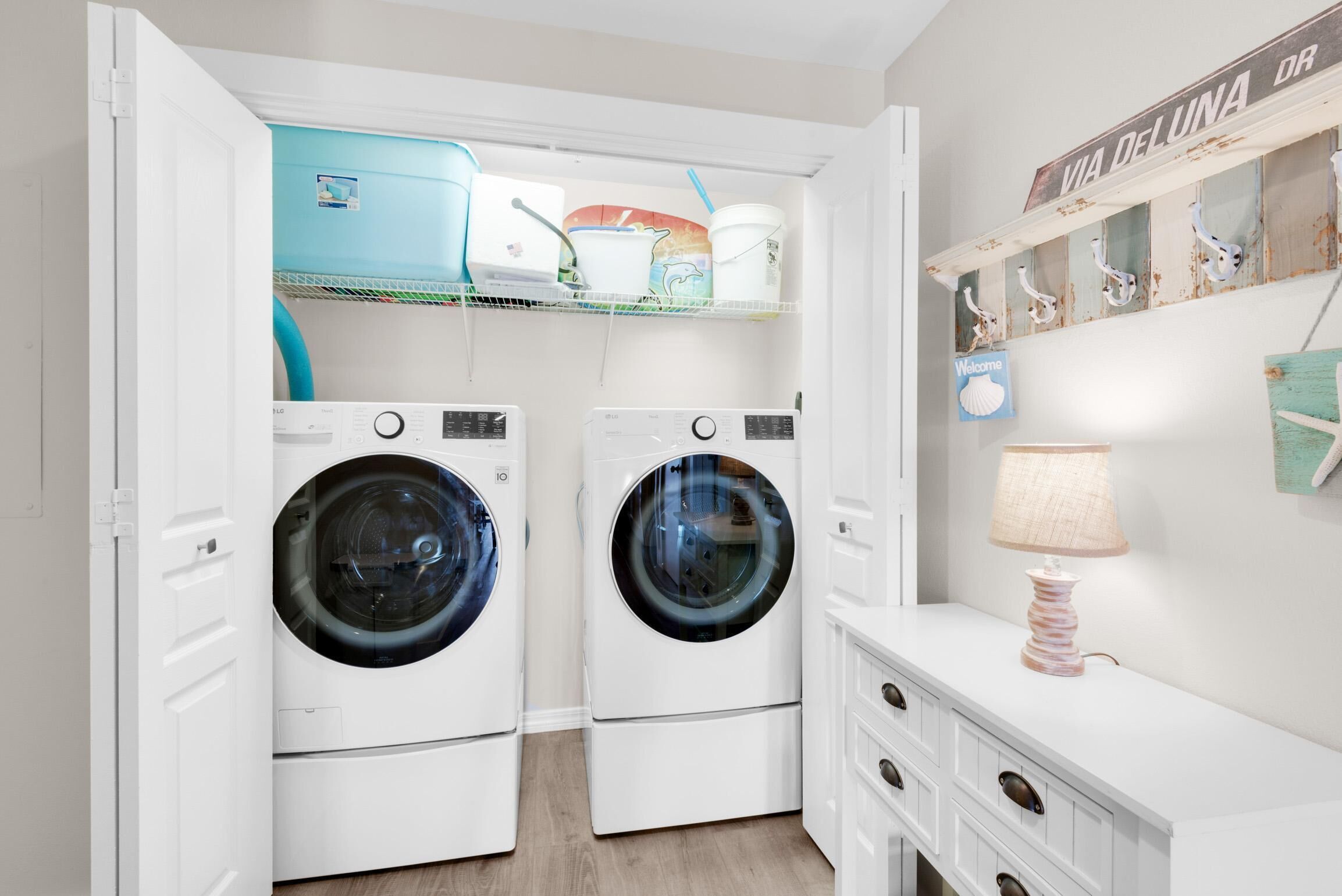 A laundry room with two front-loading washing machines, a shelf with laundry supplies, and a 'Welcome' sign.