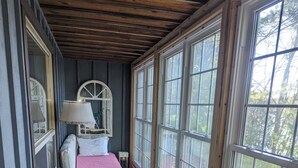 Une pièce étroite et fermée, avec un plafond en bois et de grandes fenêtres. On y trouve un lit recouvert d’une couverture rose, une petite table avec une lampe, et deux chaises en bois ornées de coussins à motifs floraux.