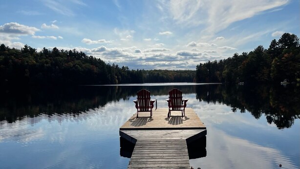 Une jetée en bois s’étendant dans une étendue d’eau calme, avec deux chaises Adirondack disposées face à l’eau, le tout entouré d’une forêt dense.