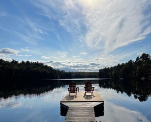 A wooden dock extending into calm water with two Adirondack chairs facing the water, surrounded by dense forest. - Fisher's Cove Sanctuary on Moon River, Muskoka (MacTier)