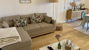 A living room with a sofa, a floor lamp, a table with a vase and remote controls, and a cabinet with decorative items. - Apartment with swimming pool - near train station (Villers-sur-Mer)