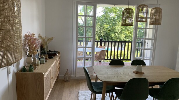 A dining area with a wooden table and chairs, a woven lamp, and a view of a garden through large windows. - Apartment with swimming pool - near train station (Villers-sur-Mer)
