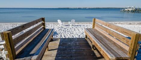 A wooden pier leading to a sandy beach with two white chairs and a wooden bench.