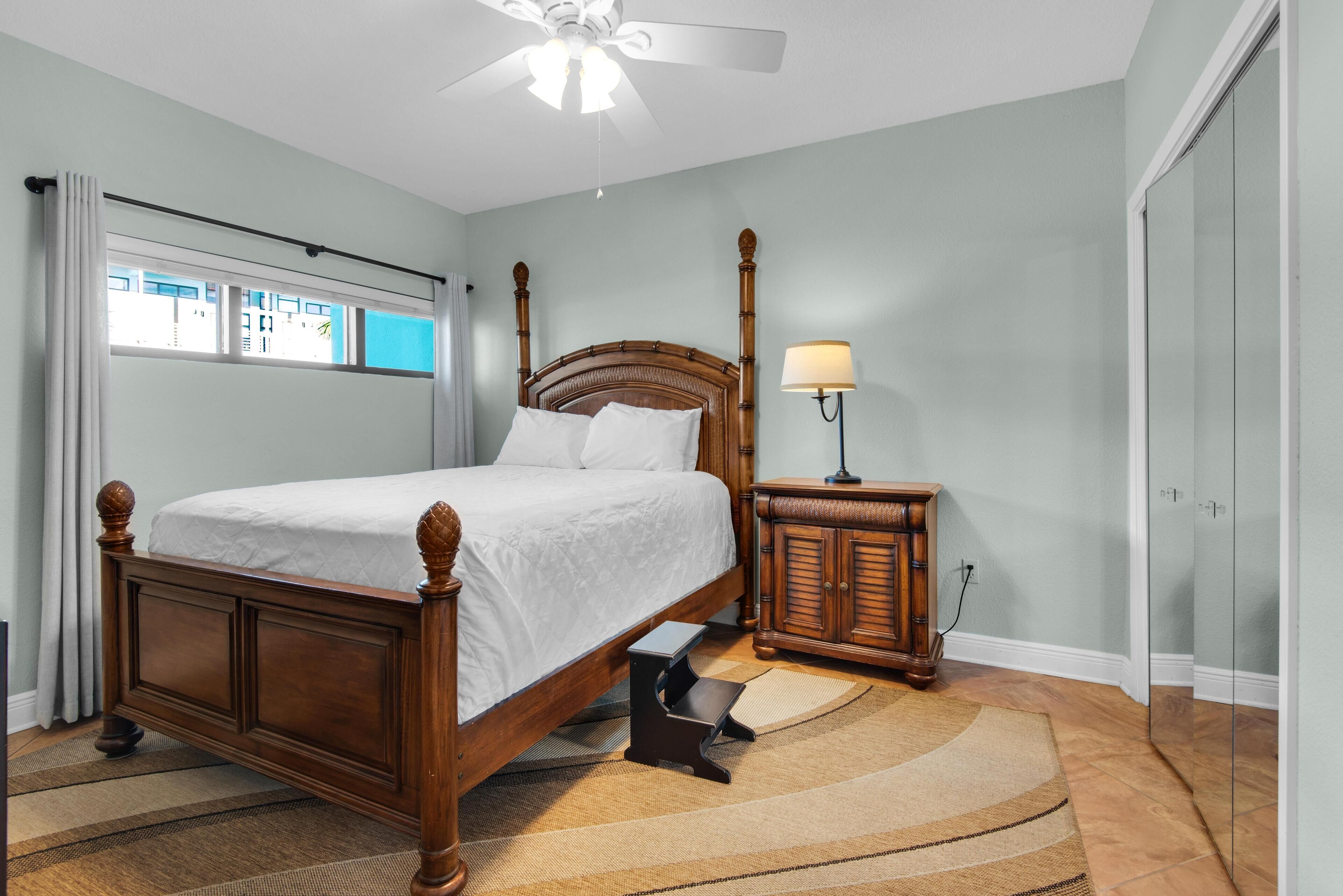A bedroom with a wooden bed, a nightstand, a lamp, a ceiling fan, and a view of the outside through a window.