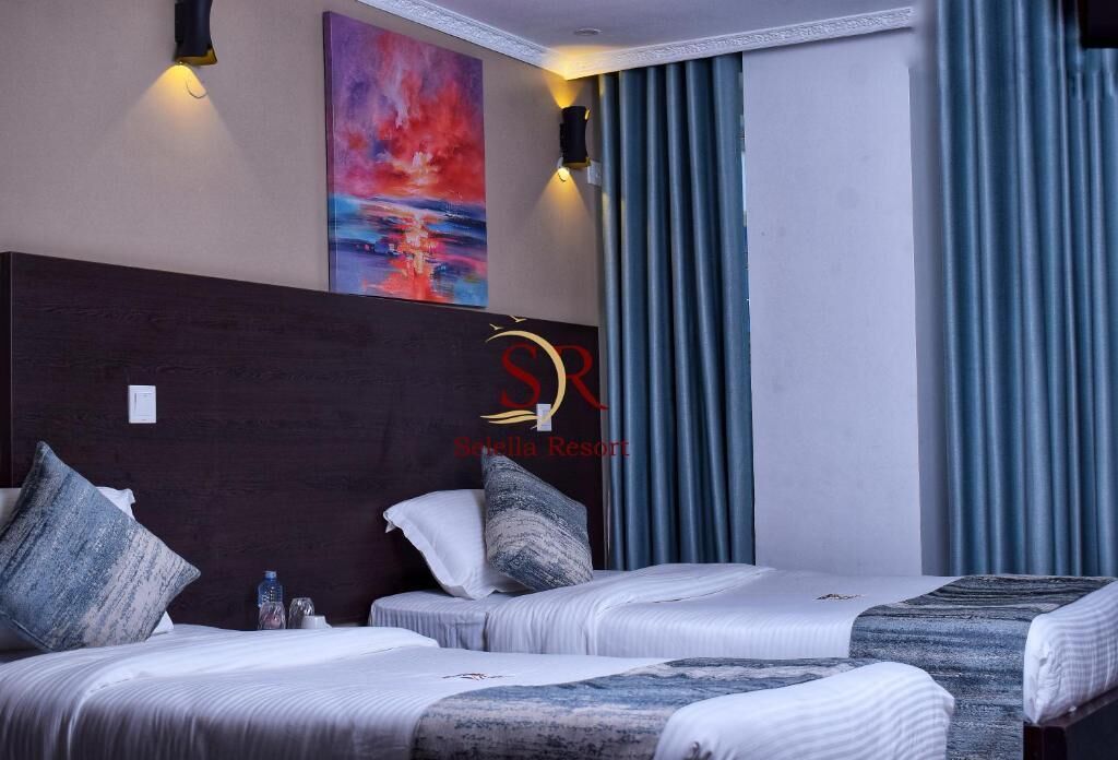 A hotel room with two beds, a large painting, and a view of the sea through curtains.