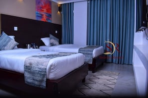 A hotel room with two beds, a headboard, a desk, and a large window with curtains. - Selella Resort (Kisumu)