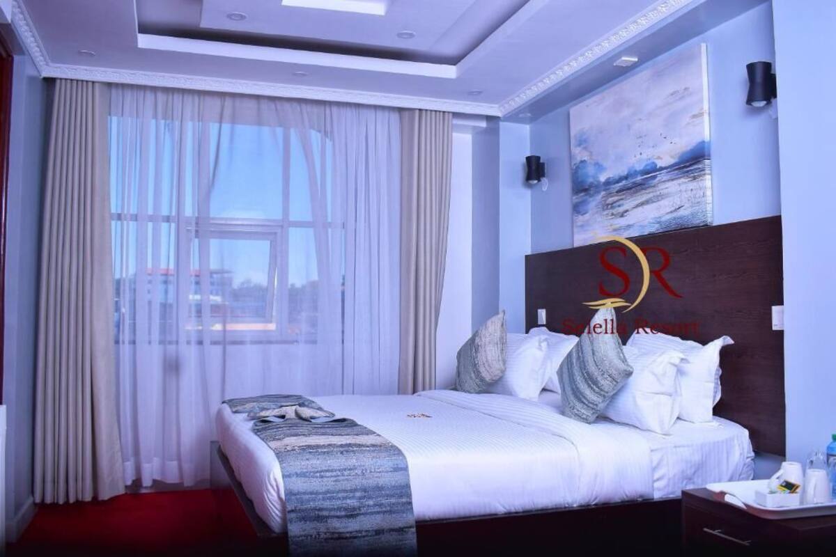 A hotel room with a large bed, a painting on the wall, and a view of the city through the window.
