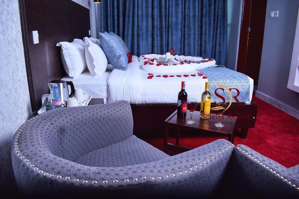 A hotel room with a bed adorned with rose petals, a small table with a bottle and glasses, and a patterned armchair.