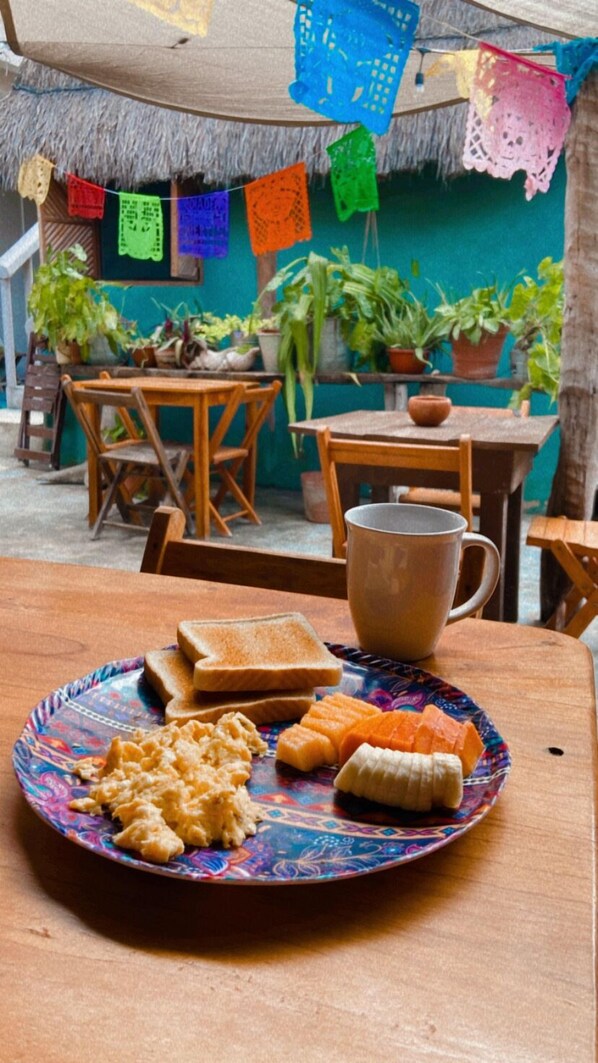 A table with a plate of food, a mug, and a thatched roof background with hanging decorations. - Barco Verde Hostel (Isla Holbox)