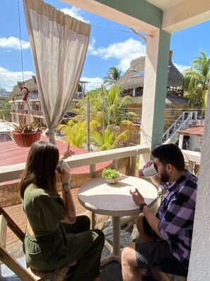 A balcony with a table and chairs, a potted plant, and a view of a thatched-roof building. - Barco Verde Hostel (Isla Holbox)