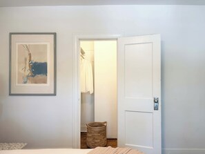 Ein Zimmer mit einem Bett, einem gerahmten abstrakten Gemälde, einer zur Garderobe führenden Tür und einem Korbwägelchen auf dem Boden.