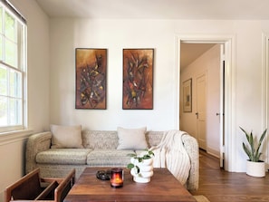 A living room with a patterned sofa, a wooden coffee table with a candle and a vase, and two abstract paintings on the wall.