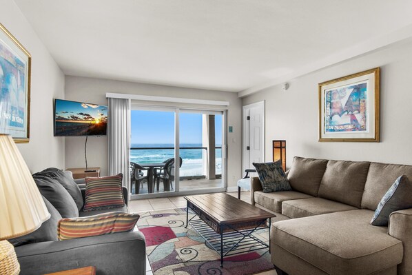 A living room with a sofa, a coffee table, and a TV. There is a view of the ocean through a sliding glass door.