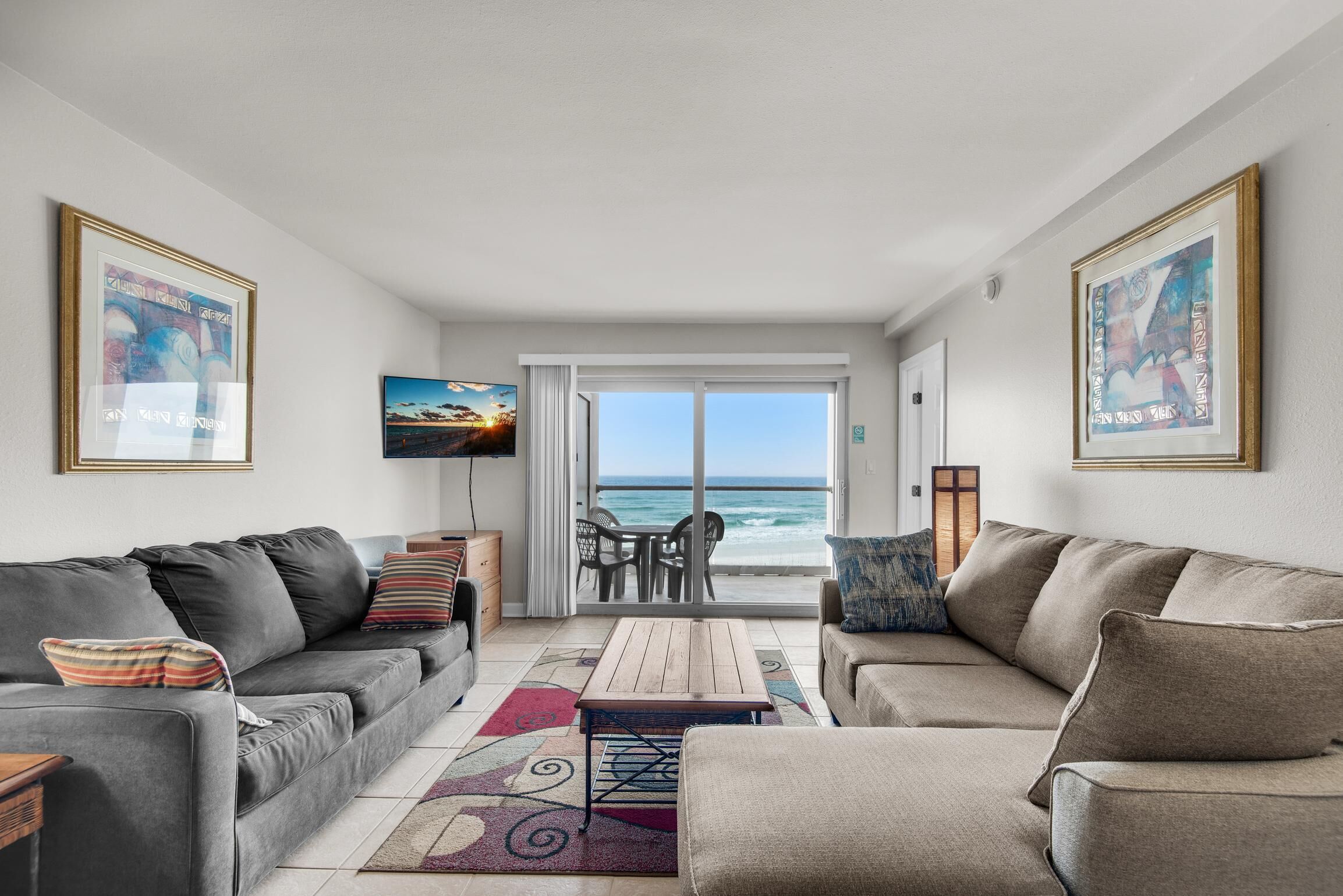 A living room with two sofas, a coffee table, and a TV. There is a view of the ocean through a glass door.