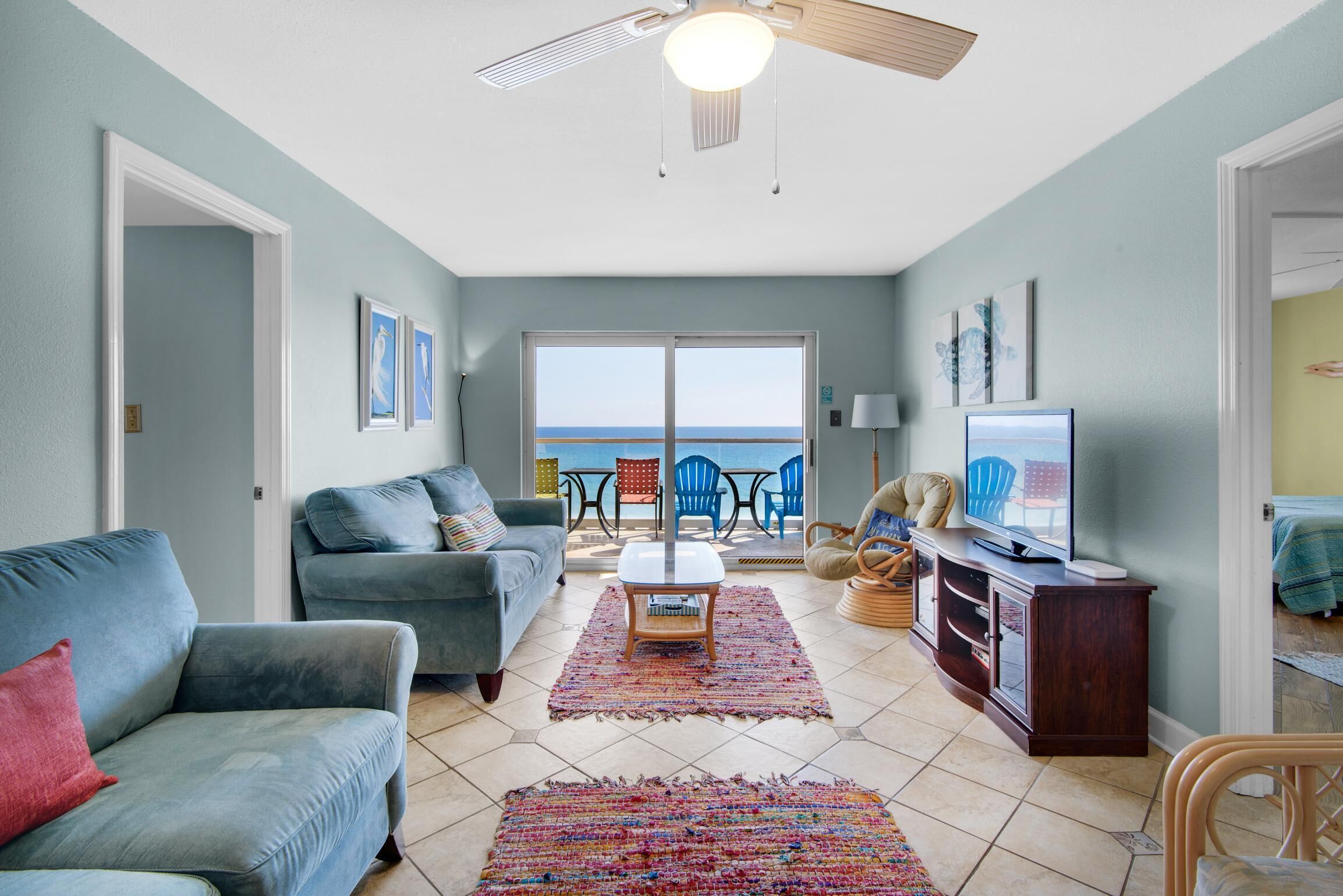 A living room with two sofas, a coffee table, and a TV on a wooden cabinet. There is a large window with a view of the ocean.