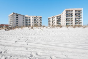 A beachfront hotel with multiple floors and balconies, situated on sandy terrain with visible footprints. - Regency Towers 702 West- Azure Pleasure (Pensacola Beach)
