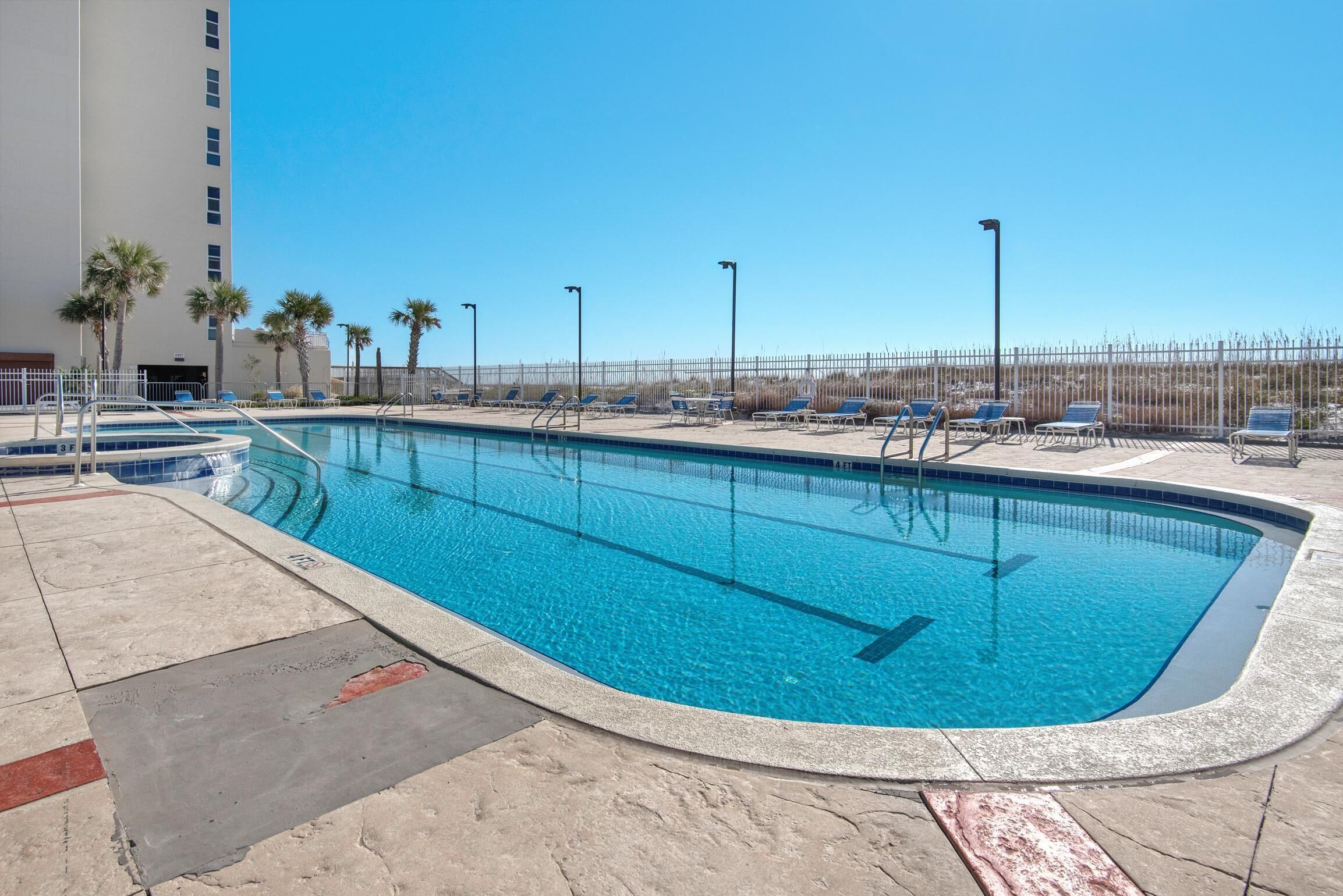 A swimming pool with clear blue water, surrounded by lounge chairs and palm trees, with a building in the background.