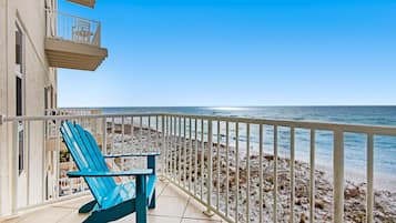 A balcony with two blue Adirondack chairs and a table, overlooking a beach with a white railing.