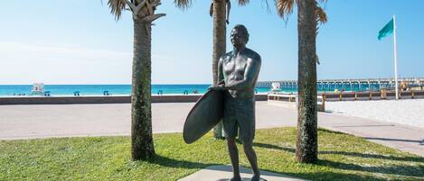 Une statue représentant un homme tenant une planche de surf sur un piédestal, entourée de palmiers, avec une plage et une jetée en arrière-plan.
