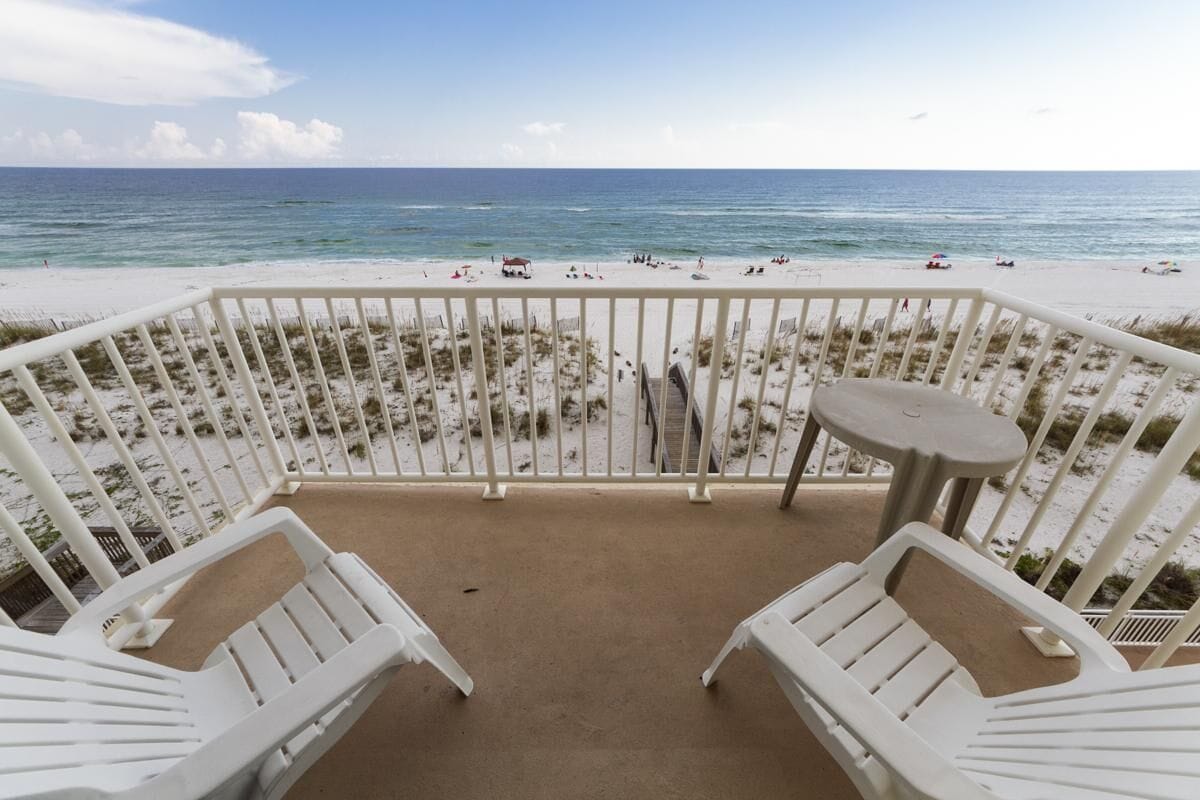 A balcony with white chairs and a small table overlooking a beach with people and umbrellas.