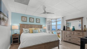 A bedroom with a bed, bedside tables, a dresser, a ceiling fan, and a large window with a view. - Sans Souci Unit 601 (Pensacola Beach)