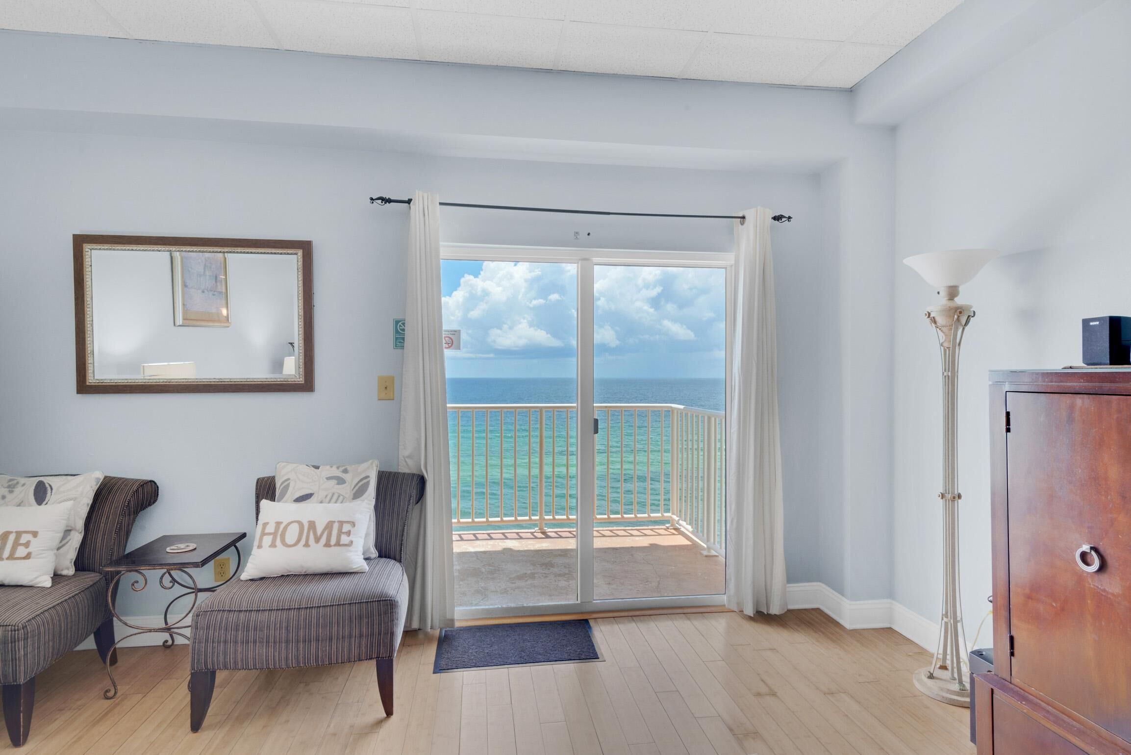 A room with a view of the sea, a balcony with a white railing, a sofa with 'HOME' pillows, a framed picture on the wall, and a tall floor lamp.