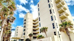 A multi-story hotel with a flat facade, balconies on each floor, and palm trees in the foreground. - Sans Souci Unit 705 (Pensacola Beach)