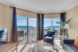 Una sala de estar con televisor, sofá, sillón, mesa y vista a la playa a través de amplias ventanas.