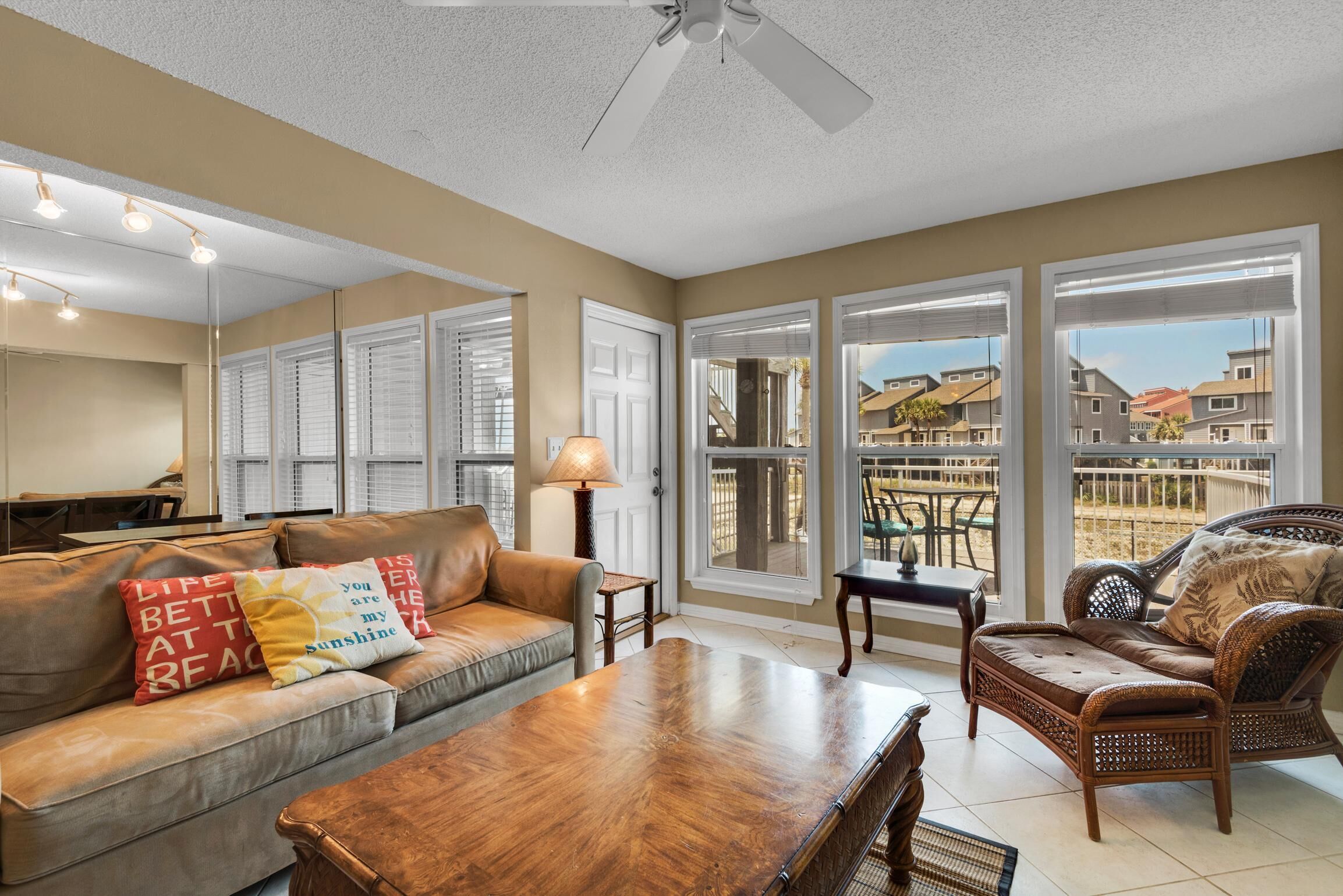 A spacious living room with a sofa, a wooden coffee table, and a wicker chair. There are large windows with blinds, a ceiling fan, and a lamp on a side table.