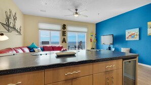 A modern kitchen with a large island, a dining area with a red couch, and a view of the sea through the windows. - Seahorse Unit M (Pensacola Beach)