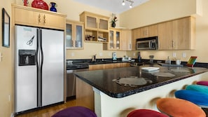 A modern kitchen with stainless steel appliances, wooden cabinets, and a central island with stools. - Seahorse Unit M (Pensacola Beach)