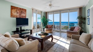 A living room with a television, a coffee table, and a sofa. There is a large window with a view of the ocean. - Emerald Dolphin Unit 510 (Pensacola Beach)